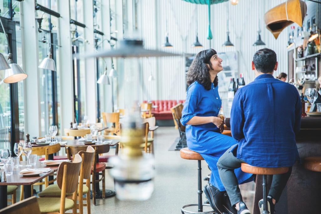 Restaurant Inside a restaurant with wooden tables and chairs along a window wall. A woman and man is sitting at a bar to the right. Blurry in the front you see an oil lamp hanging from the ceiling.