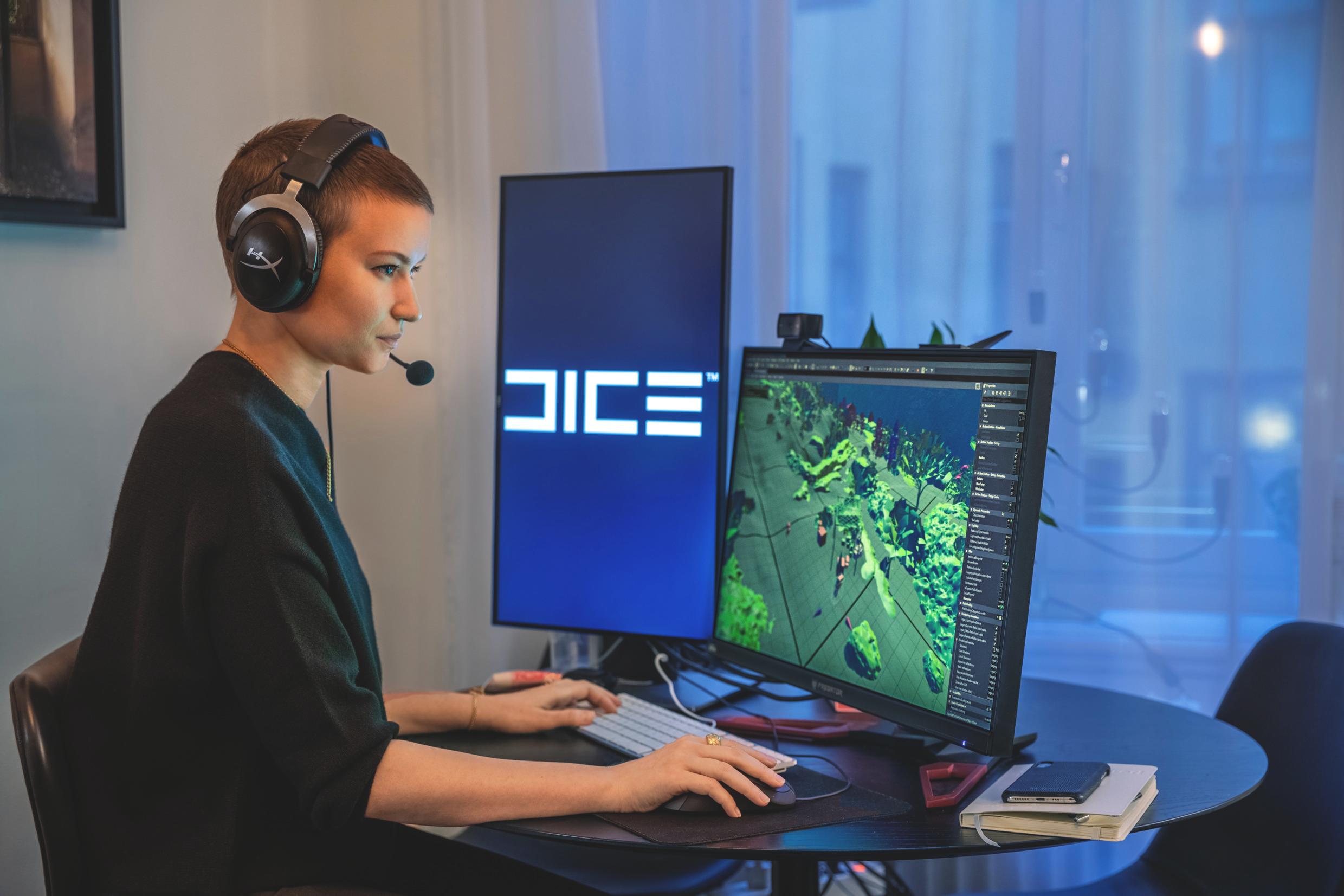 A woman sits in front of a computer with two screens. She wears a headset. On one of the screens is an ongoing game. On the other is the logo for 'Dice'.