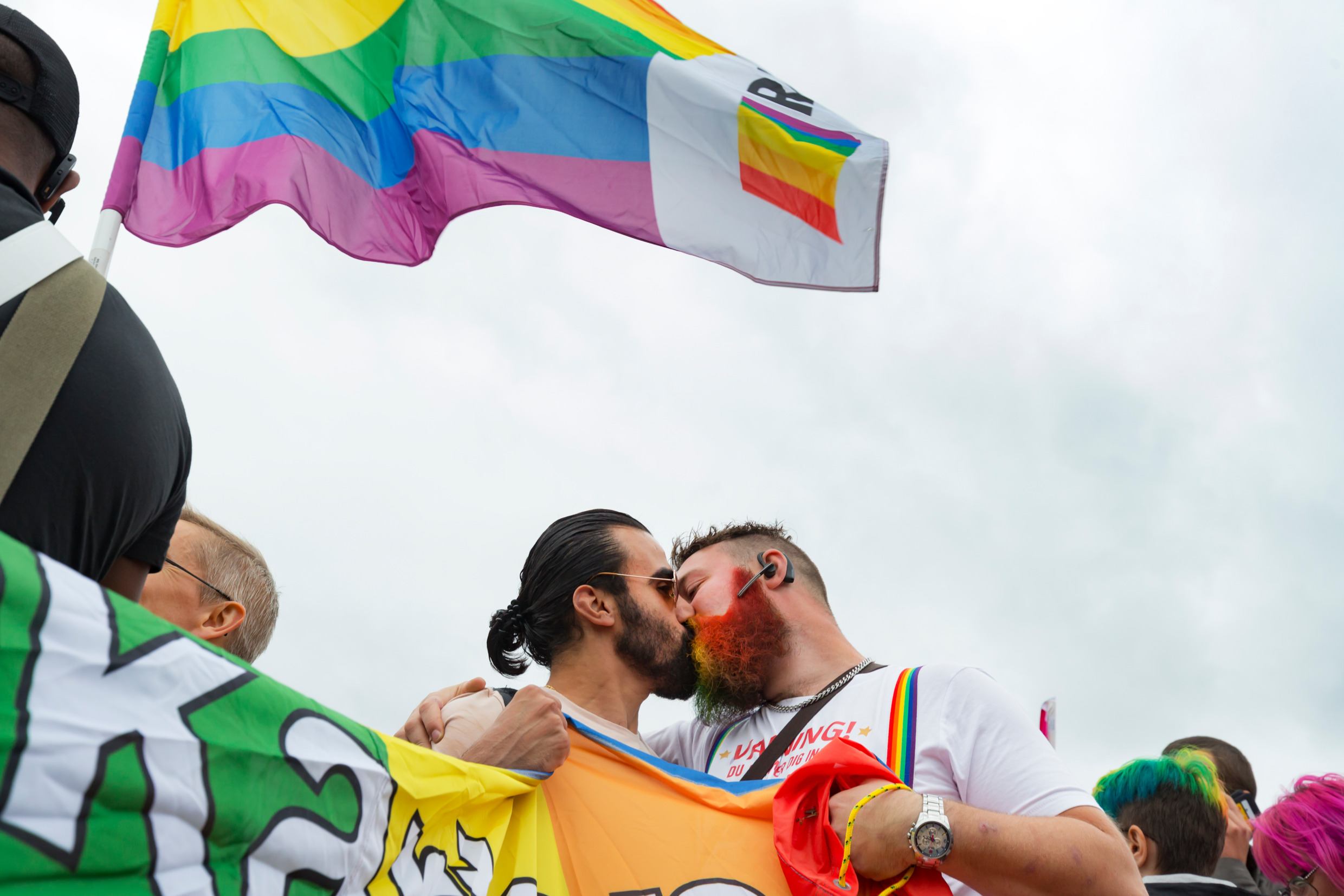 Two men kiss under a rainbow flag.