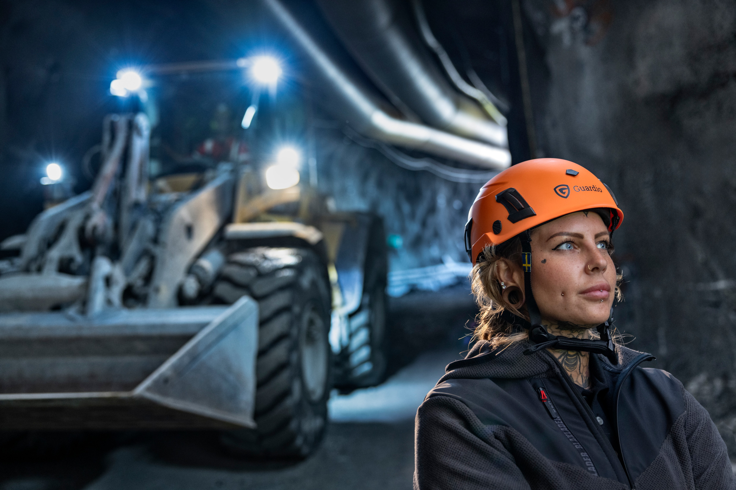 A woman with a safety helmet stands in a tunnel as a large hauler passes by in the background.